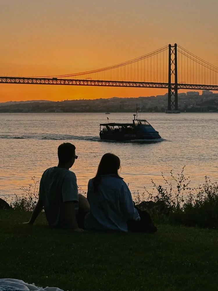 Casal sentado na grama ao pôr do sol, de costas, olhando o Tejo e a Ponte 25 de Abril em Lisboa